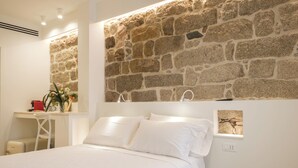 A bedroom with a stone wall, a bed with white linens, a desk with a red object, and a plant.