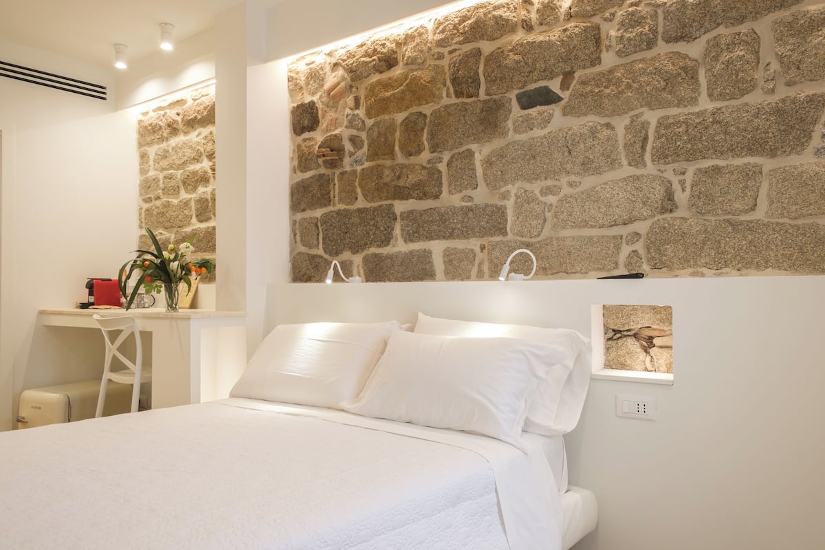 Ein Schlafzimmer mit einer Steinsäule, einem Bett mit weißen Leinen, einem Schreibtisch und einem roten Gegenstand sowie einer Pflanze.