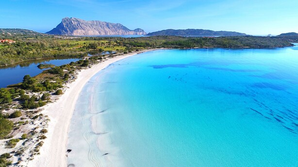 A coastal landscape with a clear blue sea, sandy beach, and a mountain in the background.