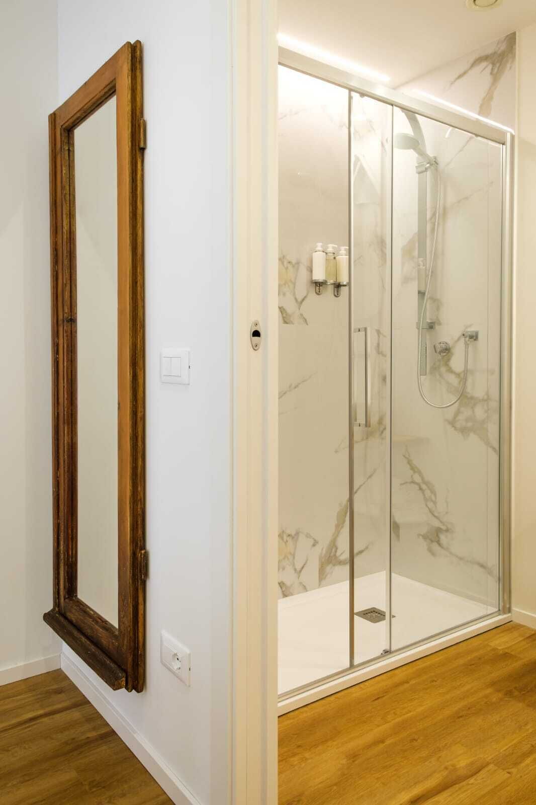 Une salle de bain dotée d’une douche à paroi de verre, d’un miroir à cadre en bois et de carreaux de marbre aux murs.