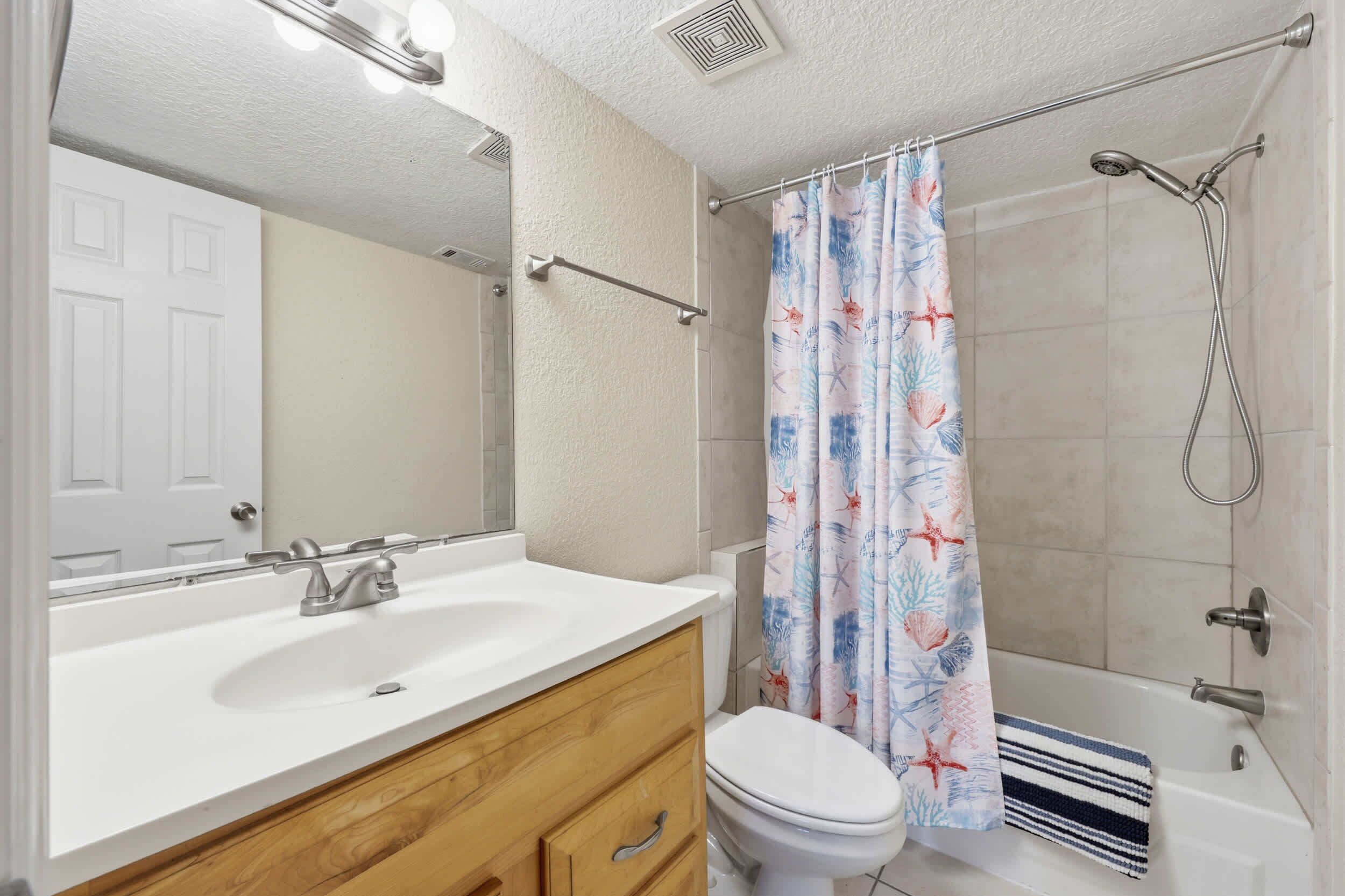 Ein Badezimmer mit weißem Waschbecken, Holz-Waschtisch, Toilette, Dusche mit Blumenvorhang und Spiegel über dem Waschbecken.