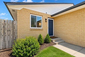 A single-story house with a brick exterior, a blue door, and a small front yard with a wooden fence and shrubs.