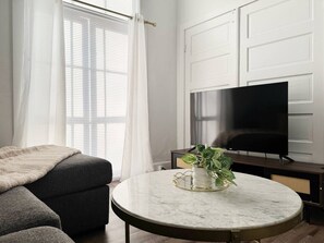 A modern living room with a dark grey sofa, a round marble coffee table with a potted plant, and a flat-screen TV on a stand. - The Harrison D - QUEEN 1BD/1BA Apt near River (Evansville)