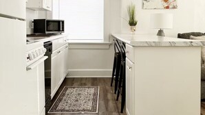 Une cuisine moderne avec des meubles blancs, un four à micro-ondes et une cuisinière. On y trouve une fenêtre avec des stores, un tapis sur le sol et un petit îlot central agrémenté d’une plante et d’une lampe.