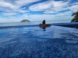 Una persona relajándose en una piscina infinita azul cristalina, con vista al océano y una pequeña isla en la distancia.