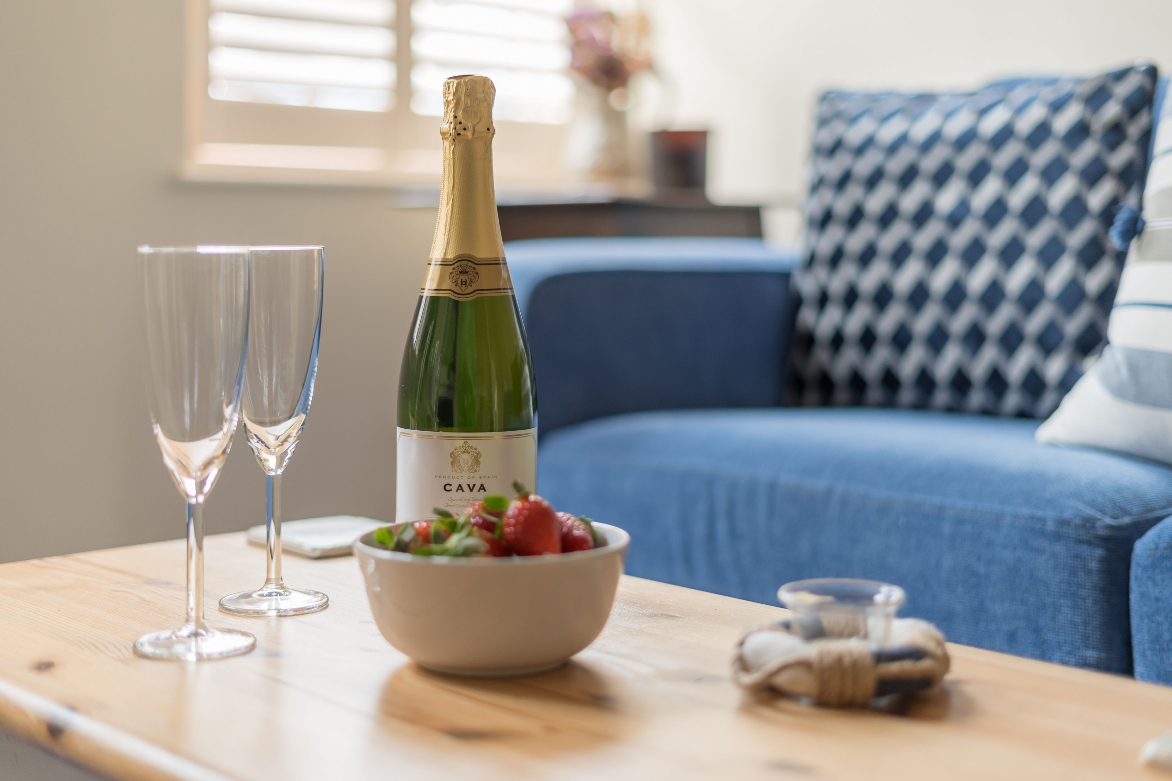 A bottle of CAVA champagne, two glasses, a bowl of strawberries, and a blue sofa with patterned cushions.