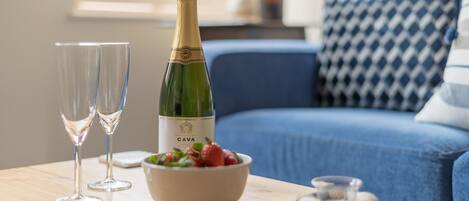 Eine Flasche CAVA Champagner, zwei Gläser, eine Schale Erdbeeren und ein blaues Sofa mit gemusterten Kissen.