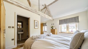 Een slaapkamer met een houten plafond, een bed met kussens, een raam met gordijn en een badkamer met wastafel en spiegel.