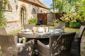 A round wicker table with chairs, a stone building with arched windows, and a wooden fence in a garden setting. - 3 Marshland Barns - sleeps 8 guests  in 4 bedrooms (Titchwell)