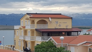 Ein zweistöckiges Gebäude mit Balkonen und einem Schild mit der Aufschrift 'Villa Bado' vor einer Berglandschaft und einem Gewässer.