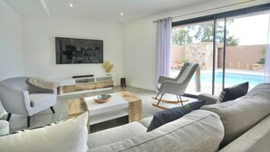 A modern living room with a sofa, armchairs, a coffee table, and a TV mounted on the wall. There is a large window with a view of a swimming pool and outdoor seating. - Villa 10 people with swimming pool - Lecci (Lecci)