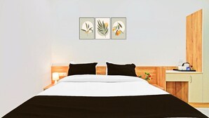 A hotel room with a bed, two black pillows, a nightstand with a lamp, a wooden desk with a cup and a book, and three framed leaf artworks on the wall.
