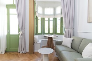 A room with a green door, a round table, a sofa, and a bay window with curtains. - Ogigou Suite (Athens)