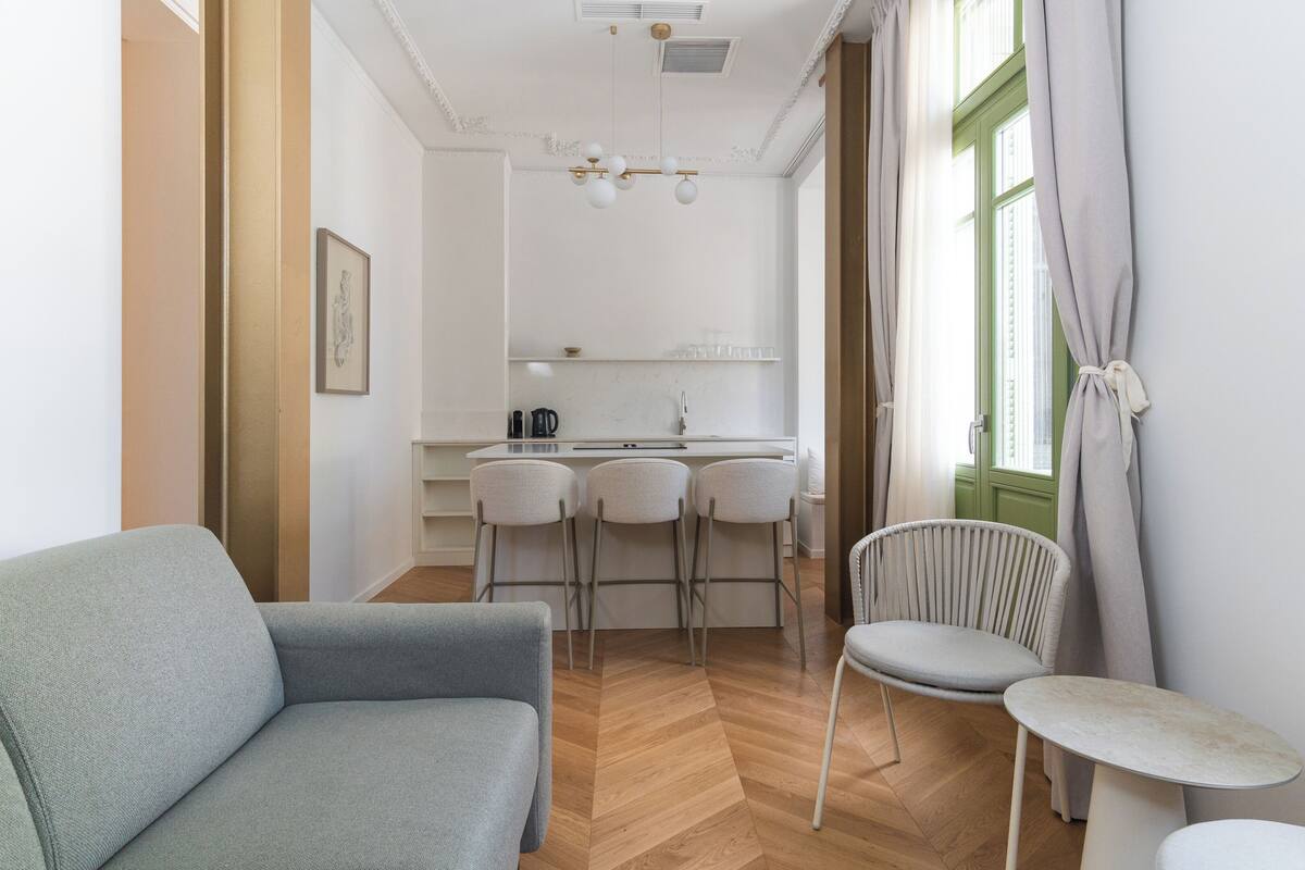 A modern living room with a sofa and chairs, a small table, and a kitchen area with a bar stool.