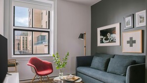 Una sala de estar moderna con un sofá gris, una silla roja, una mesita con una planta y un ventanal con vista a la ciudad.