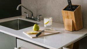 Una encimera de cocina con un fregadero, una tabla de cortar con una pera y un cuchillo, un vaso de agua con rodajas de limón y un bloque de cuchillos.