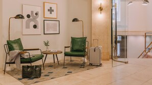 Un vestíbulo de hotel con dos sillones de cuero verde, una mesita con una planta, un bolso negro y una maleta. Se observan obras de arte enmarcadas en la pared y una partición de vidrio con una puerta.