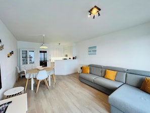 Ein modernes Wohnzimmer mit grauem Sofa, einem Essbereich mit Tisch und Stühlen sowie einer Küche im Hintergrund.