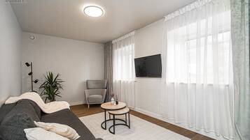 Un salon avec un canapé gris, une table basse ronde, un fauteuil gris, une plante en pot, un lampadaire et un téléviseur fixé au mur.