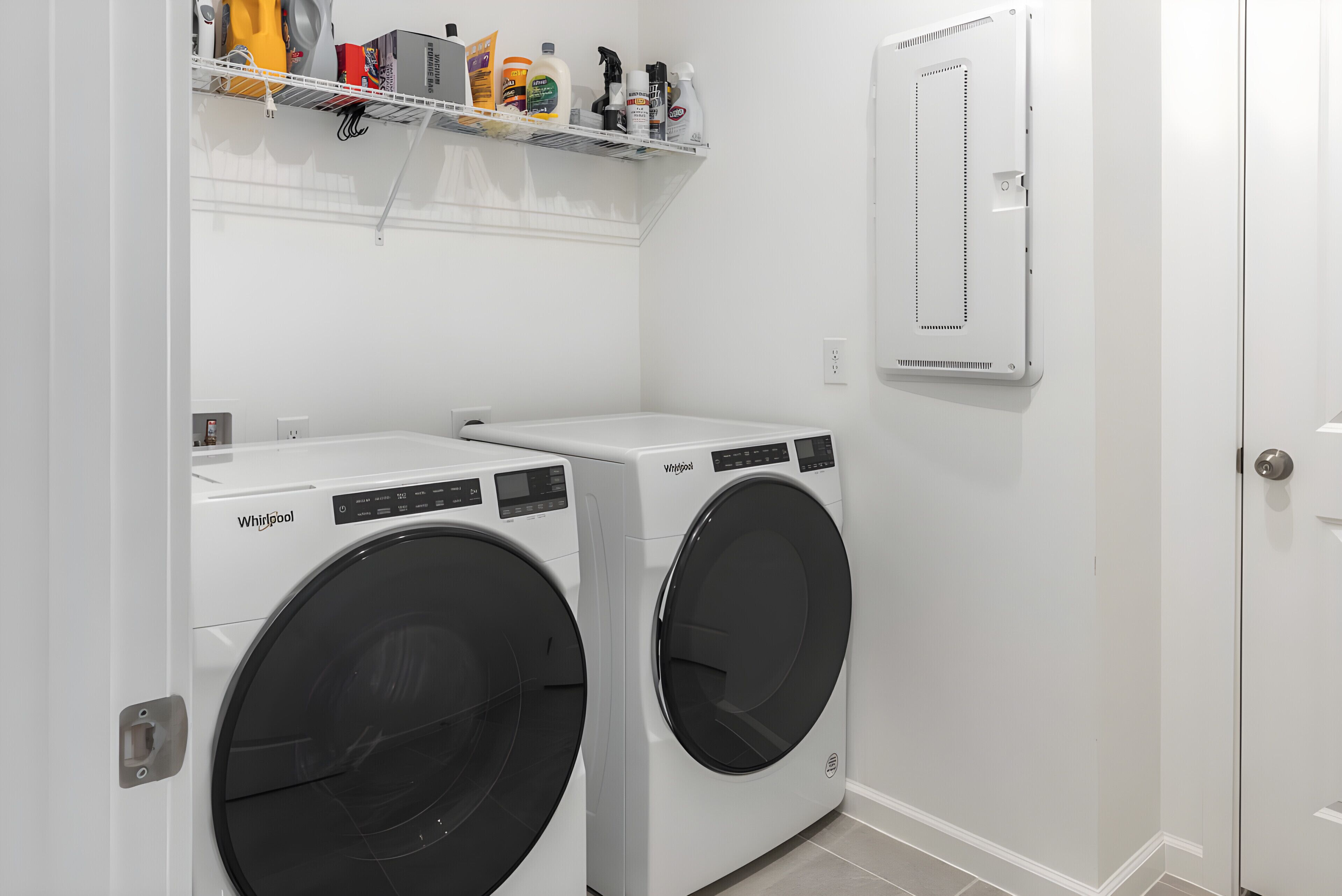 Ein Waschraum mit zwei Whirlpool Frontlader-Waschmaschinen, einem Regal mit Reinigungsmitteln und einer weißen Tür mit Spiegel.