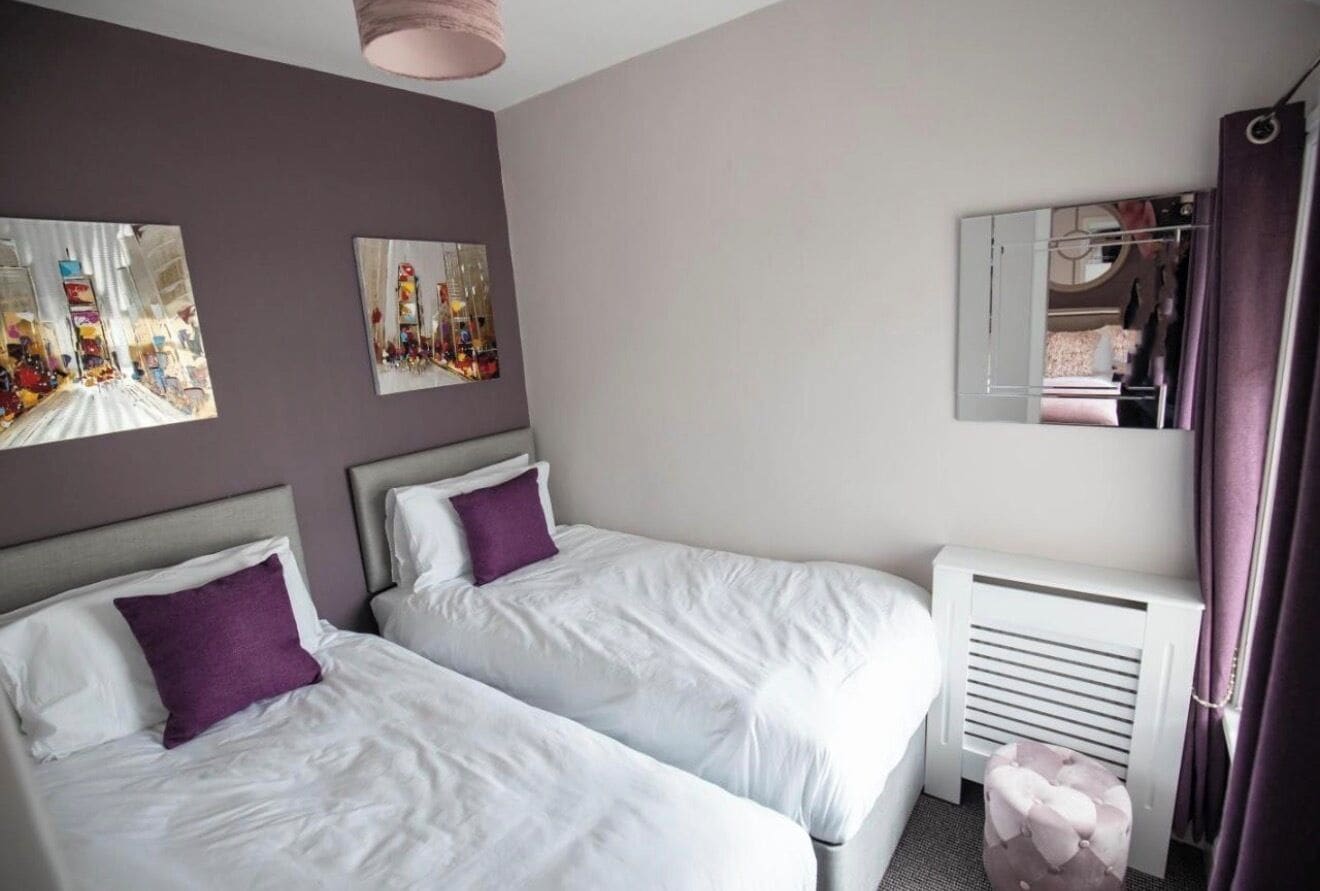 A hotel room with two beds, a radiator, a mirror, and artwork on the wall.