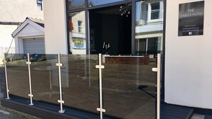 Une entrée d’hôtel moderne avec une façade en verre, un portail de sécurité en métal et une enseigne indiquant le nom de l’hôtel et ses coordonnées.