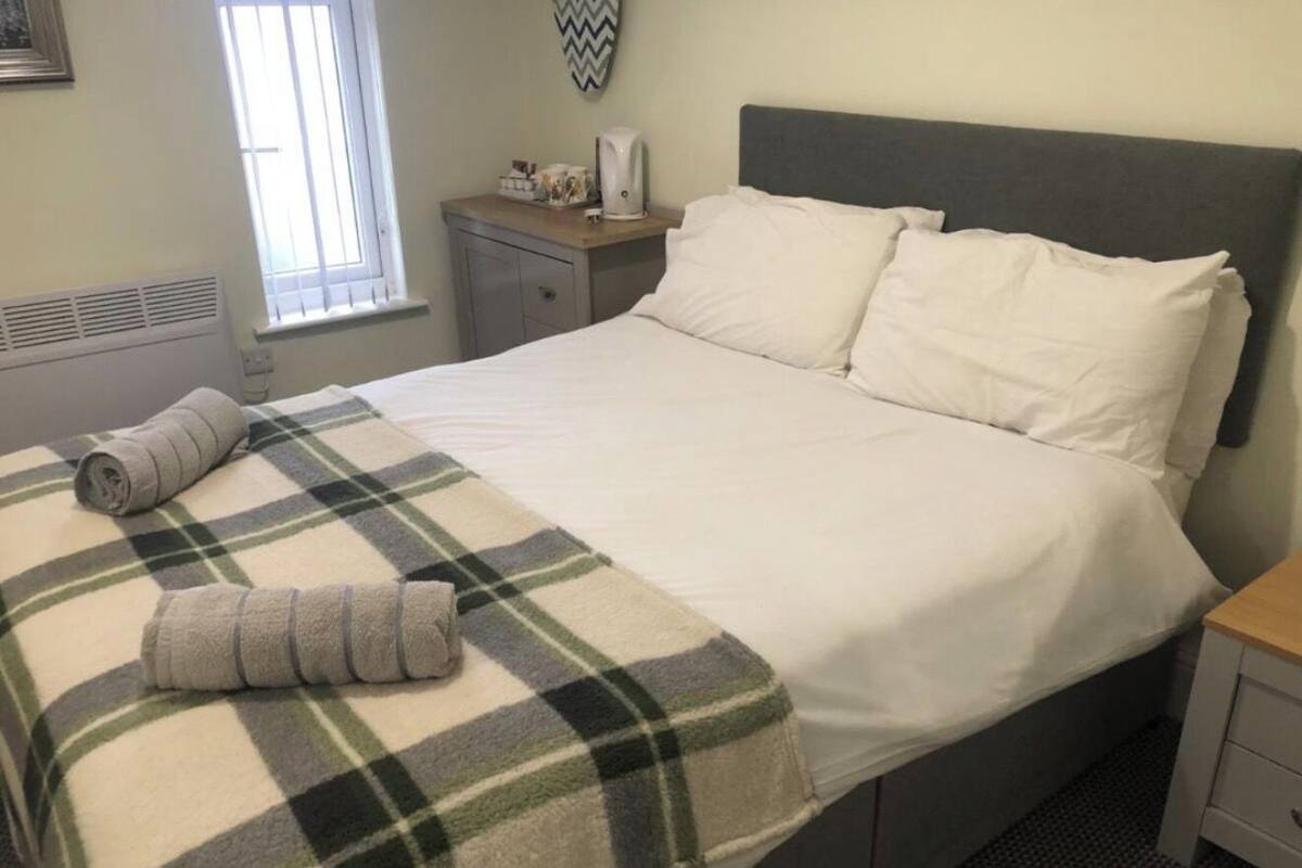 Un lit bien fait, avec une couverture à carreaux et deux oreillers, une table de chevet avec une lampe et une tasse, et une fenêtre avec des stores.