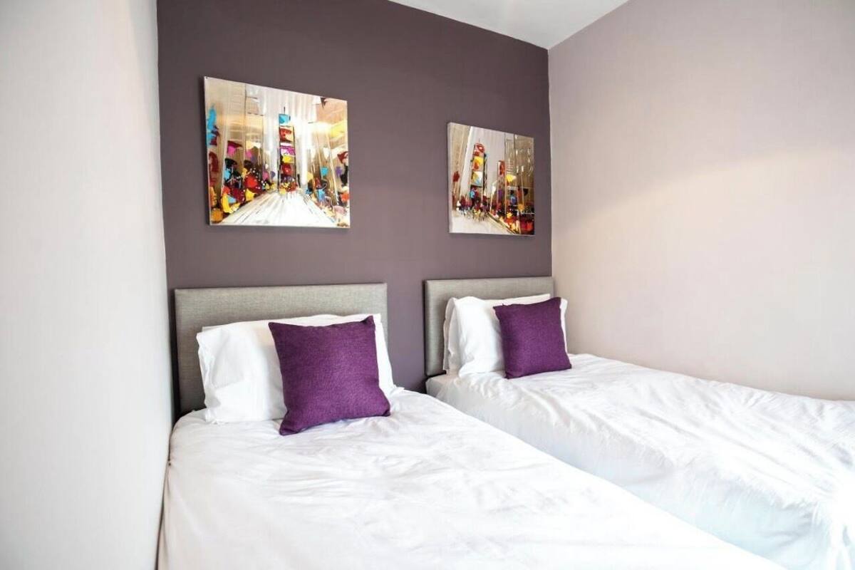 Une chambre d’hôtel avec deux lits, chacun agrémenté de draps blancs et d’oreillers violets. Les murs sont peints dans une couleur claire, et deux grandes peintures sont accrochées au-dessus des lits.
