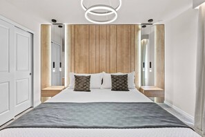 Ein modernes Schlafzimmer mit einem hölzernen Kopfteil, einem großen Bett mit grauer Decke sowie Nachttischen mit Lampen.