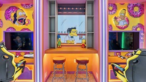Quarto com parede temática dos Simpsons, área de bar com banquetas e uma tela de televisão.