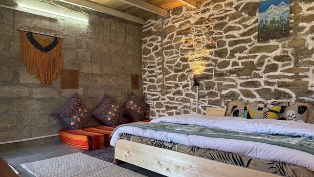Una habitación rústica con pared de piedra, cama de madera, almohadones de colores y un tapiz tejido.
