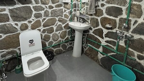 A bathroom with a stone wall, a white sink, a toilet, and a green bucket. - Enjoyable 1-bedroom bed & breakfast in charming Naggar (Naggar)