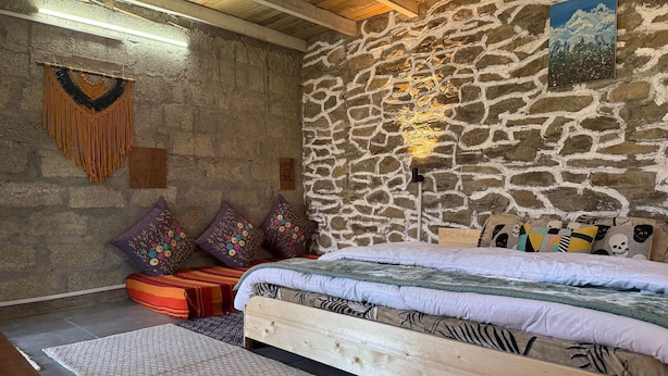 A rustic room with a stone wall, a wooden bed, colorful pillows, and a woven rug. - Enjoyable 1-bedroom bed & breakfast in charming Naggar (Naggar)