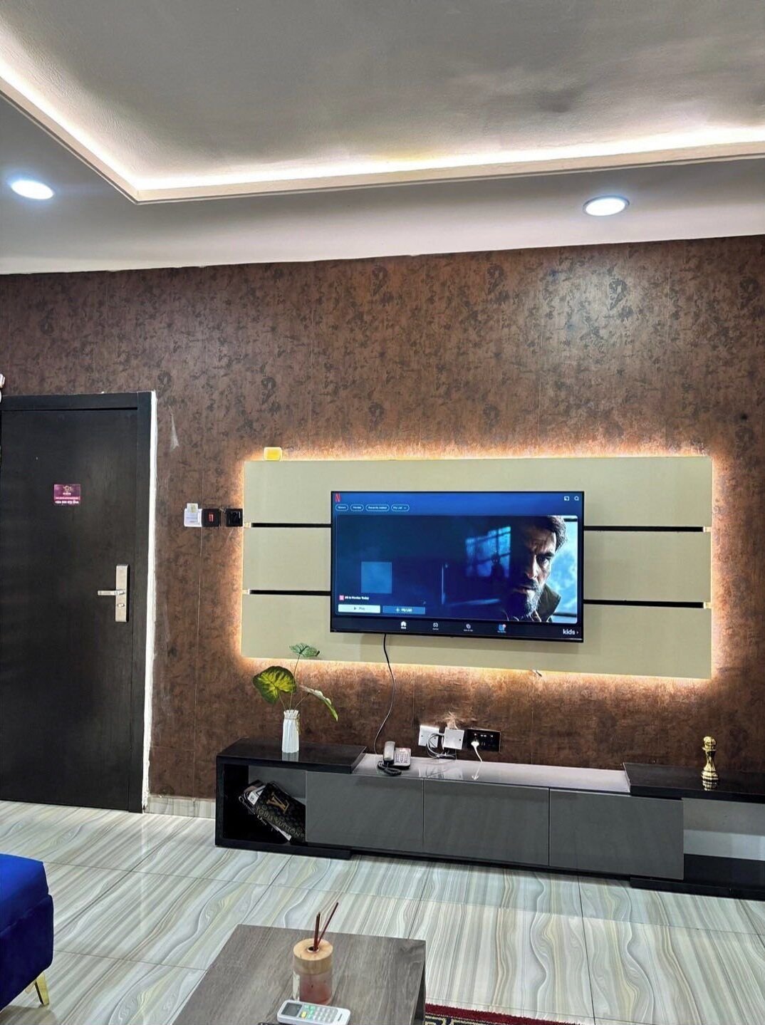 A modern living room with a flat-screen TV mounted on a textured wall, a dark wooden door, and a blue sofa.