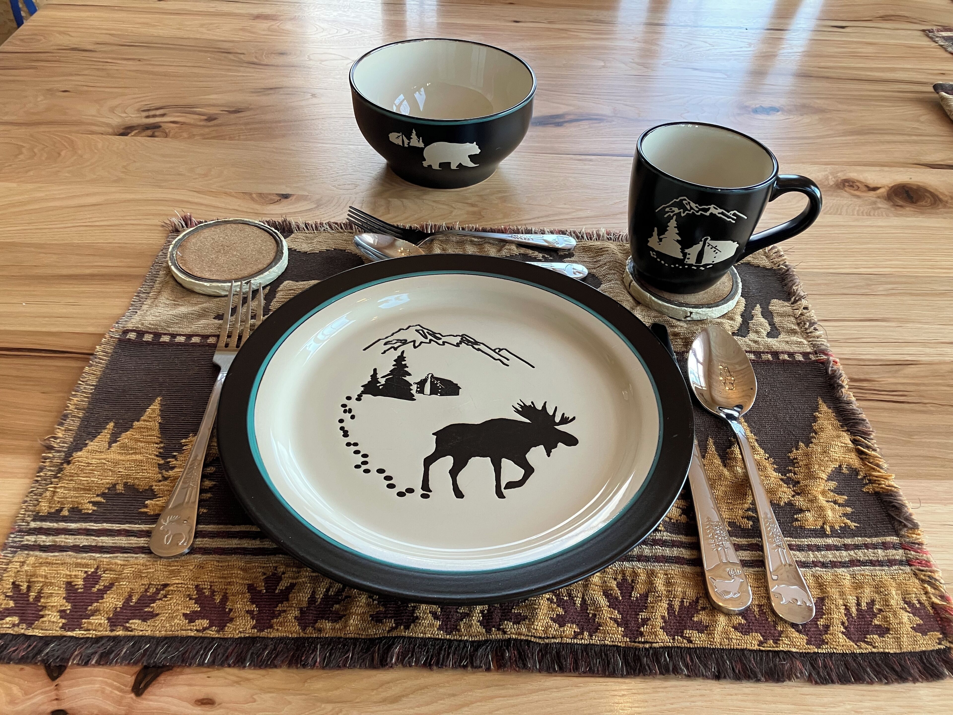 A table set with a plate featuring a moose and mountains, a black mug with a mountain design, and a black bowl with a bear silhouette.