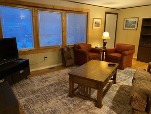 A living room with a wooden coffee table, two orange armchairs, a sofa, a TV on a stand, and a lamp on a side table. - 2 BR house, close to national parks and ski hill (Golden)