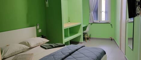 A bedroom with a bed, a shelf unit, a window with curtains, and a wall-mounted TV.