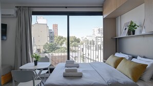 A modern hotel room with a large bed, a small table with chairs, a potted plant, and a view of the cityscape through large windows. - MOT Rentals + Design SohoHaus '407' - 3 pax Full Amenities (Buenos Aires)