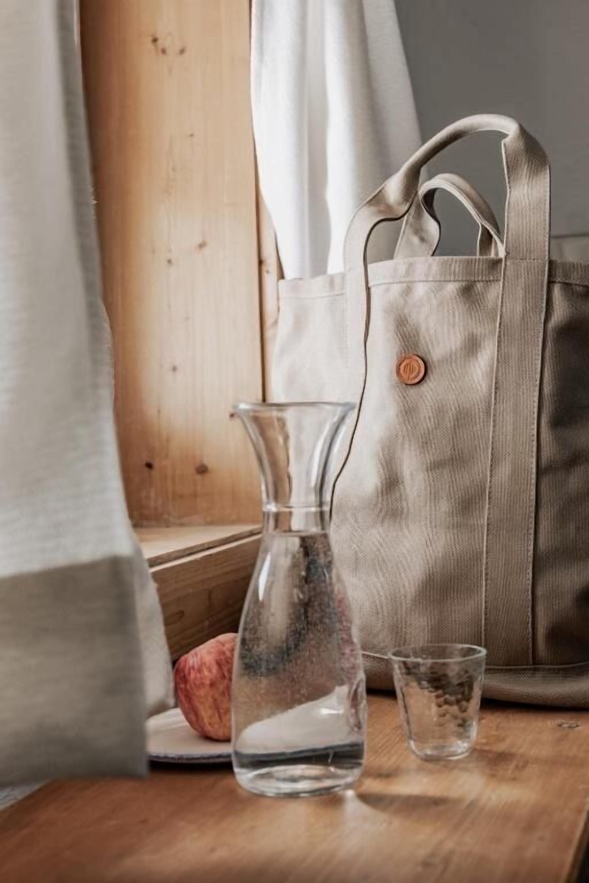A wooden table with a clear glass bottle, a small glass, and a beige bag.