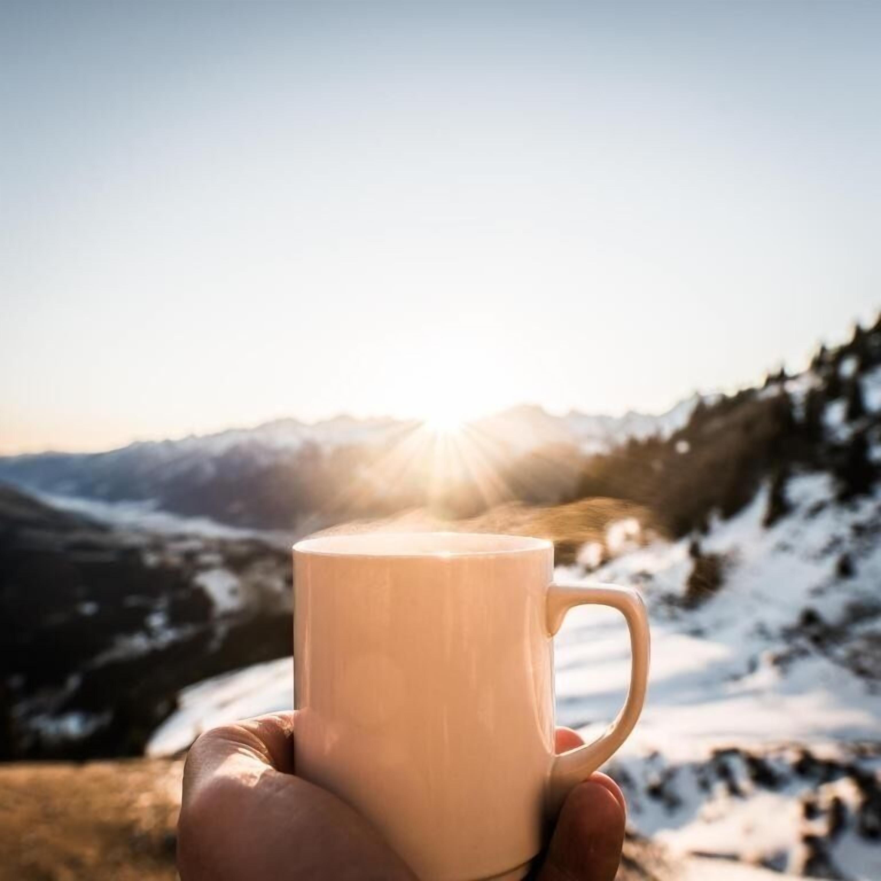 A hand holding a mug with steam rising from it, set against a mountainous landscape with the sun shining in the background.
