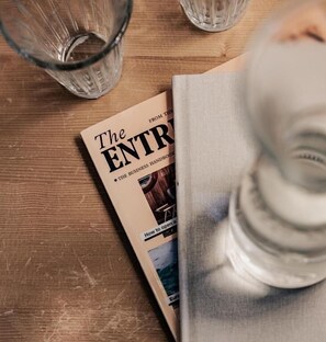 Una rivista intitolata 'The ENTRY' su un tavolo di legno, con due bicchieri trasparenti e una bottiglia d'acqua.
