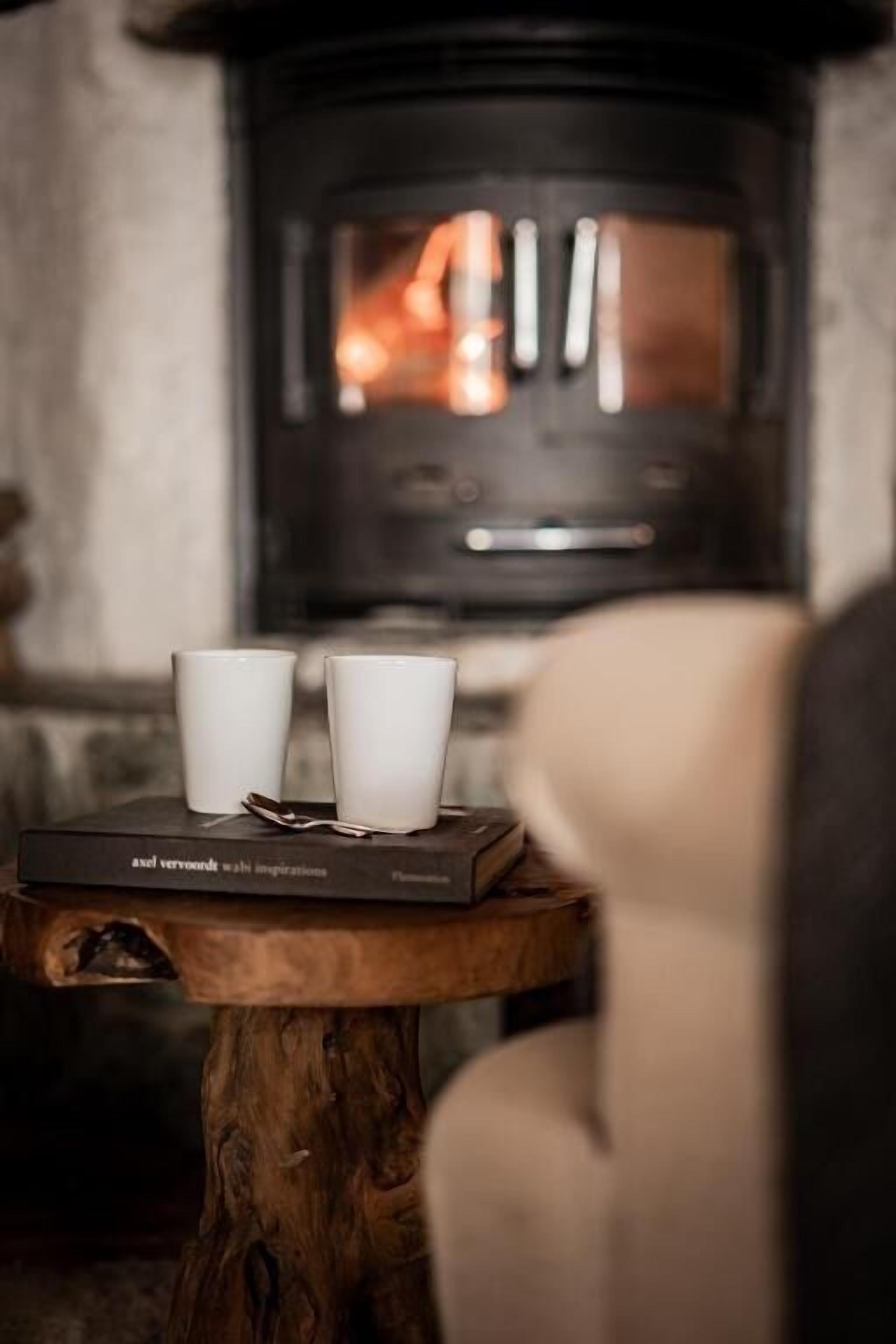 Quarto acolhedor com lareira, duas chávenas brancas sobre uma mesa de madeira, e um livro intitulado 'Ael Verwoerdt: Ali Inspirations'.