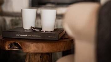 Quarto acolhedor com lareira, duas chávenas brancas sobre uma mesa de madeira, e um livro intitulado 'Ael Verwoerdt: Ali Inspirations'.
