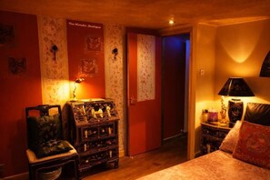 Een kamer met een bed, een houten kastje met beeldjes, een rode deur en een bloemen behang muur.