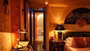 A hotel room with a bed, a nightstand with a lamp, a wall-mounted fan, and a door leading to another room. - Oriental Suite (Swansea)