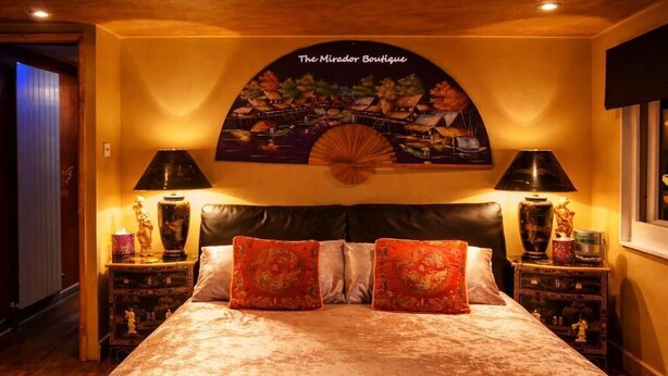 A bedroom with a large bed, two nightstands, and a wall-mounted artwork depicting a tropical scene. - Oriental Suite (Swansea)