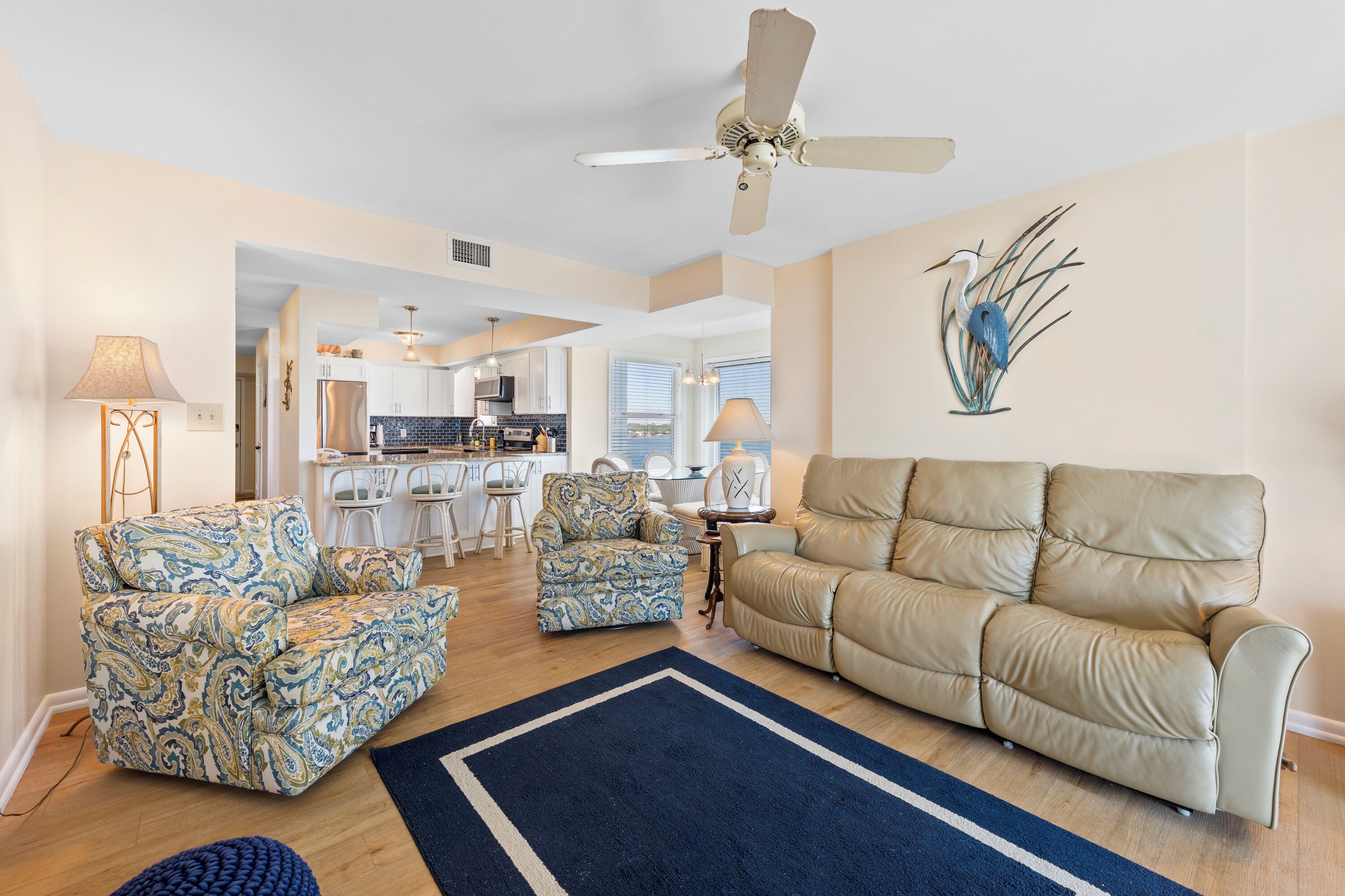 A spacious living room with a beige sofa, patterned armchairs, and a blue rug. A ceiling fan and a wall-mounted bird sculpture are visible.