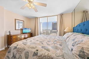 A hotel room with a large bed, a ceiling fan, a television, and a view of the city through the window. - Compass Point 510 (Gulf Shores)