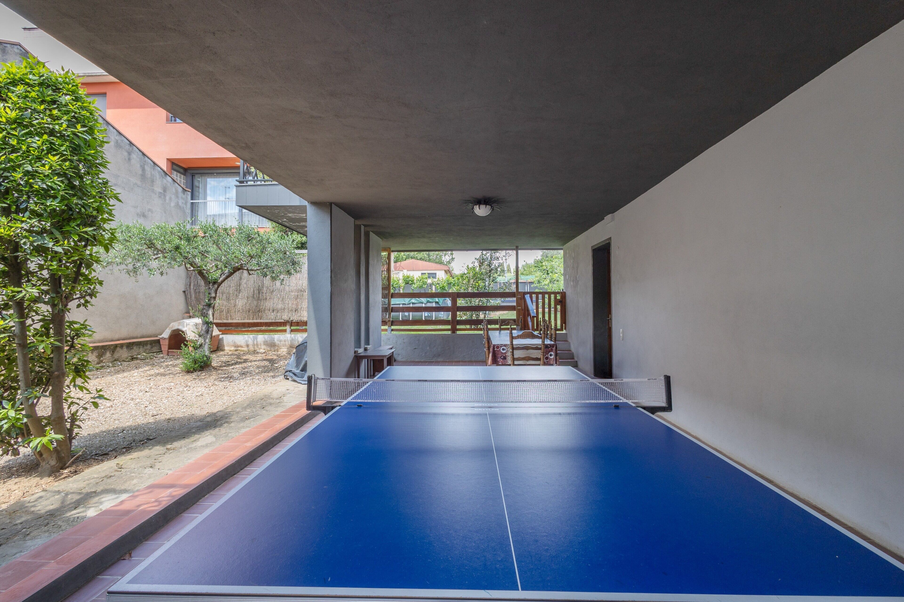 Ein Pingpongtisch in einem Außenbereich mit Betonplafond, einer kleinen Sitzecke und Blick auf ein Gebäude im Hintergrund.