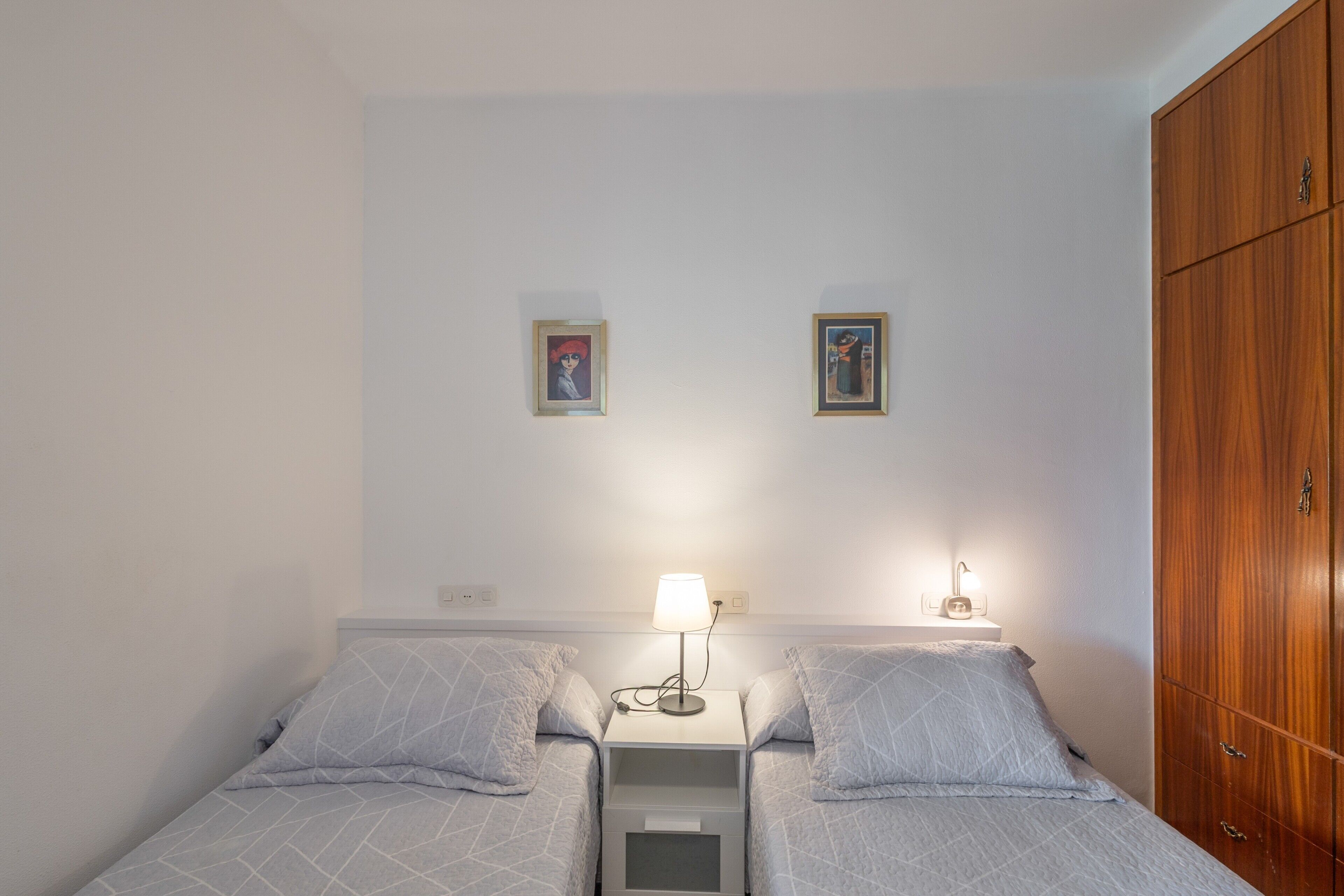 A bedroom with two beds, a nightstand with a lamp, and a wardrobe.